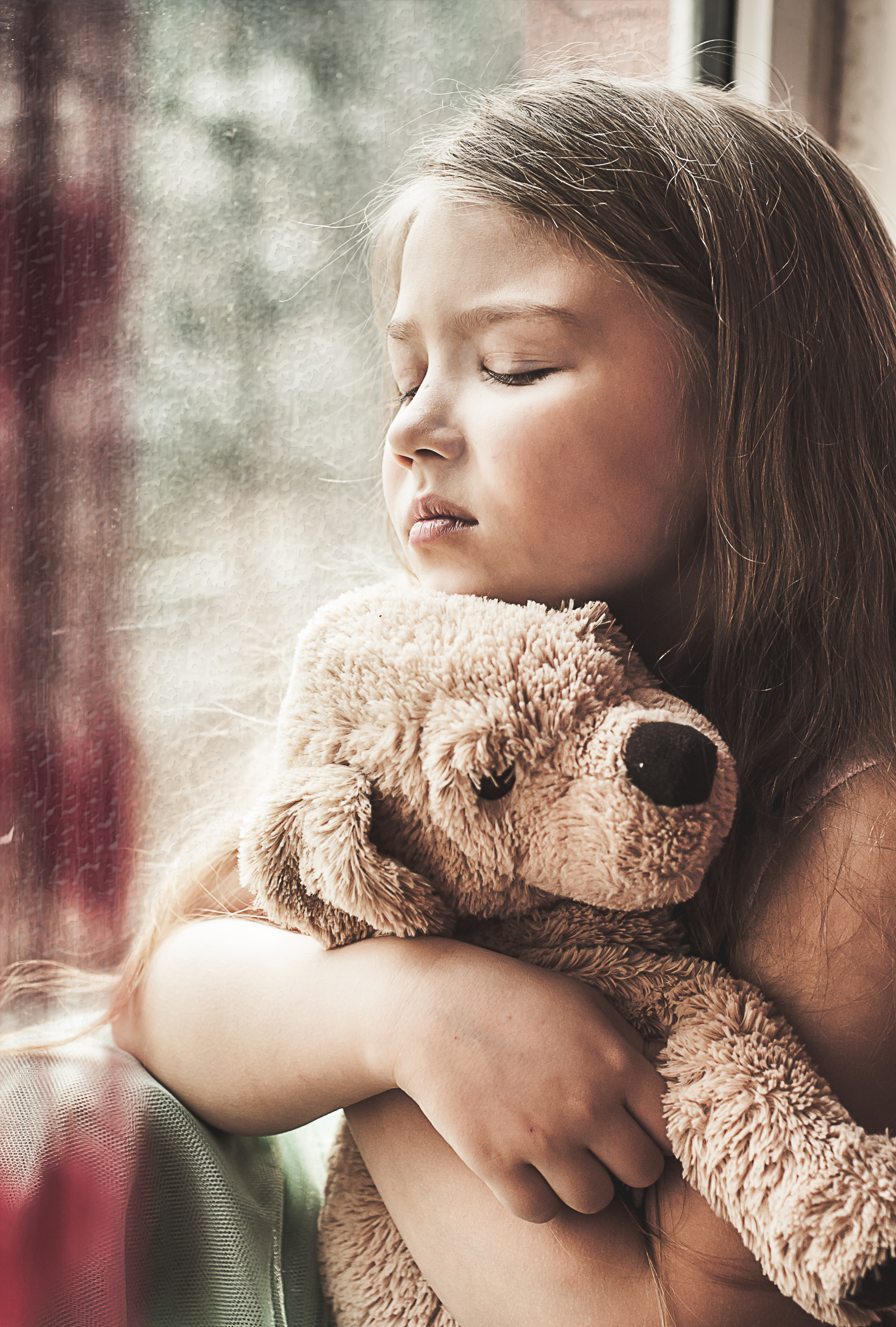 Little beautiful sad unhappy girl with puppy toys sits on windowsill
